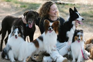 Woman sitting on ground surrounding by several dog breeds